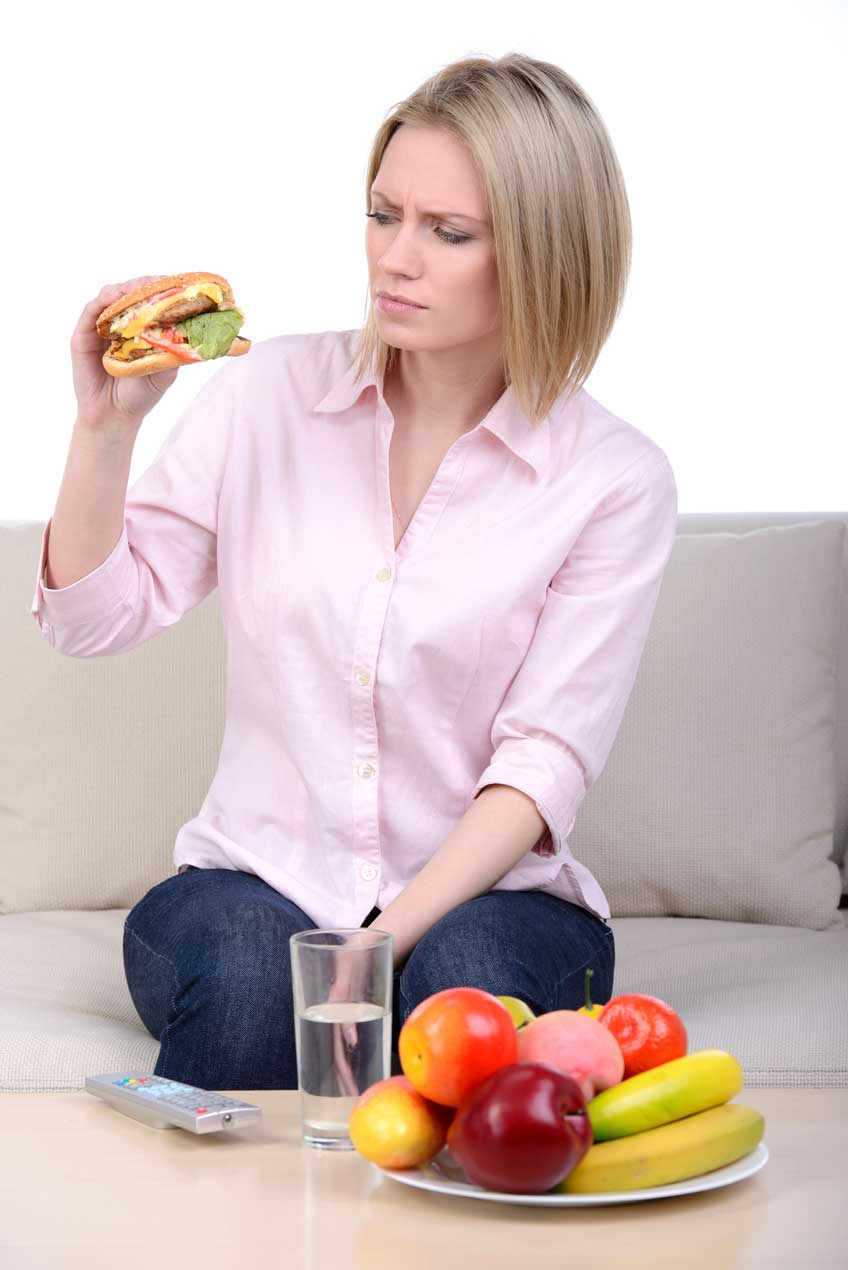 Grávida com sanduíche na mão, e frutas sobre a mesa à sua frente.